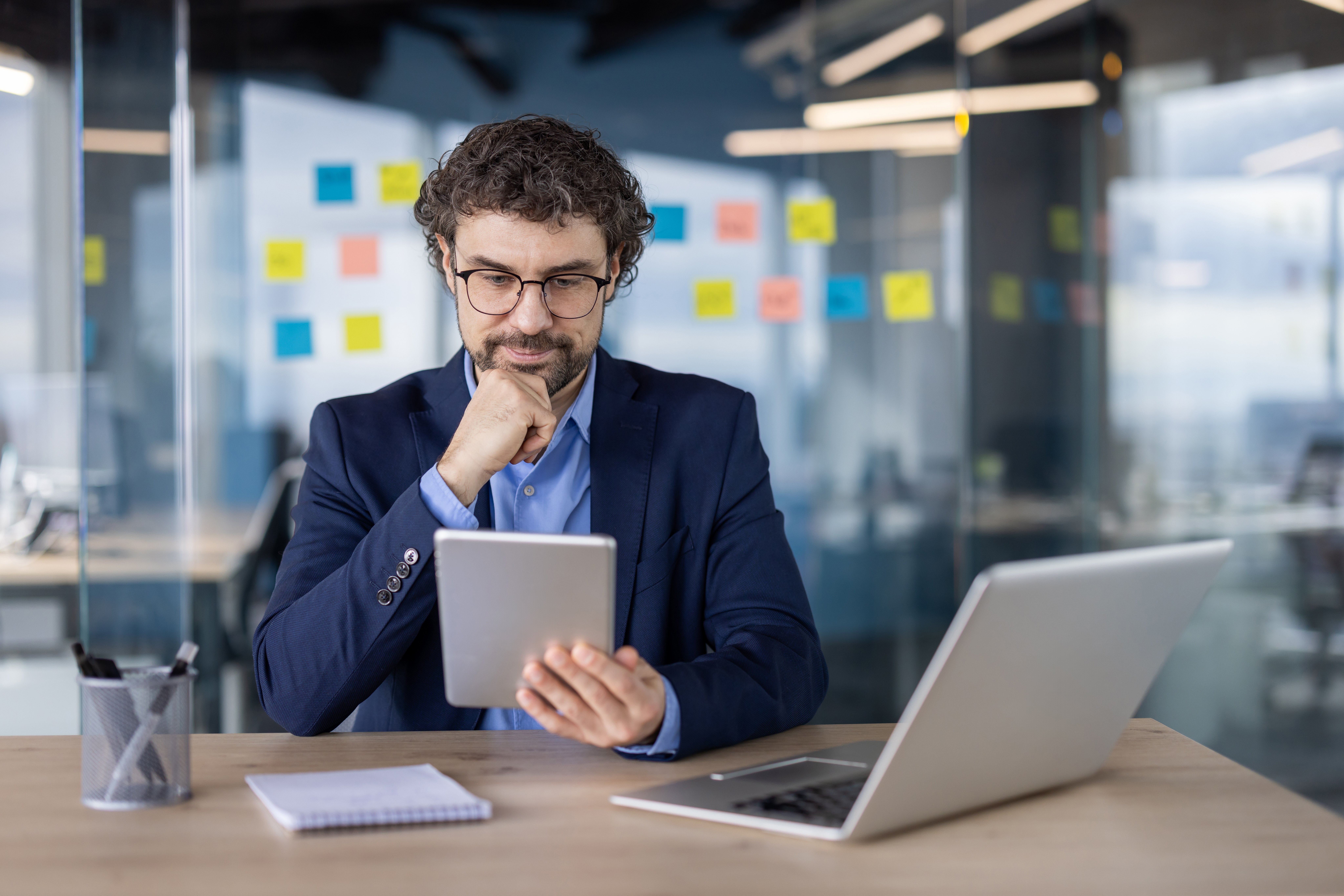 Businessman in modern office using digital tablet while thinking and working on laptop