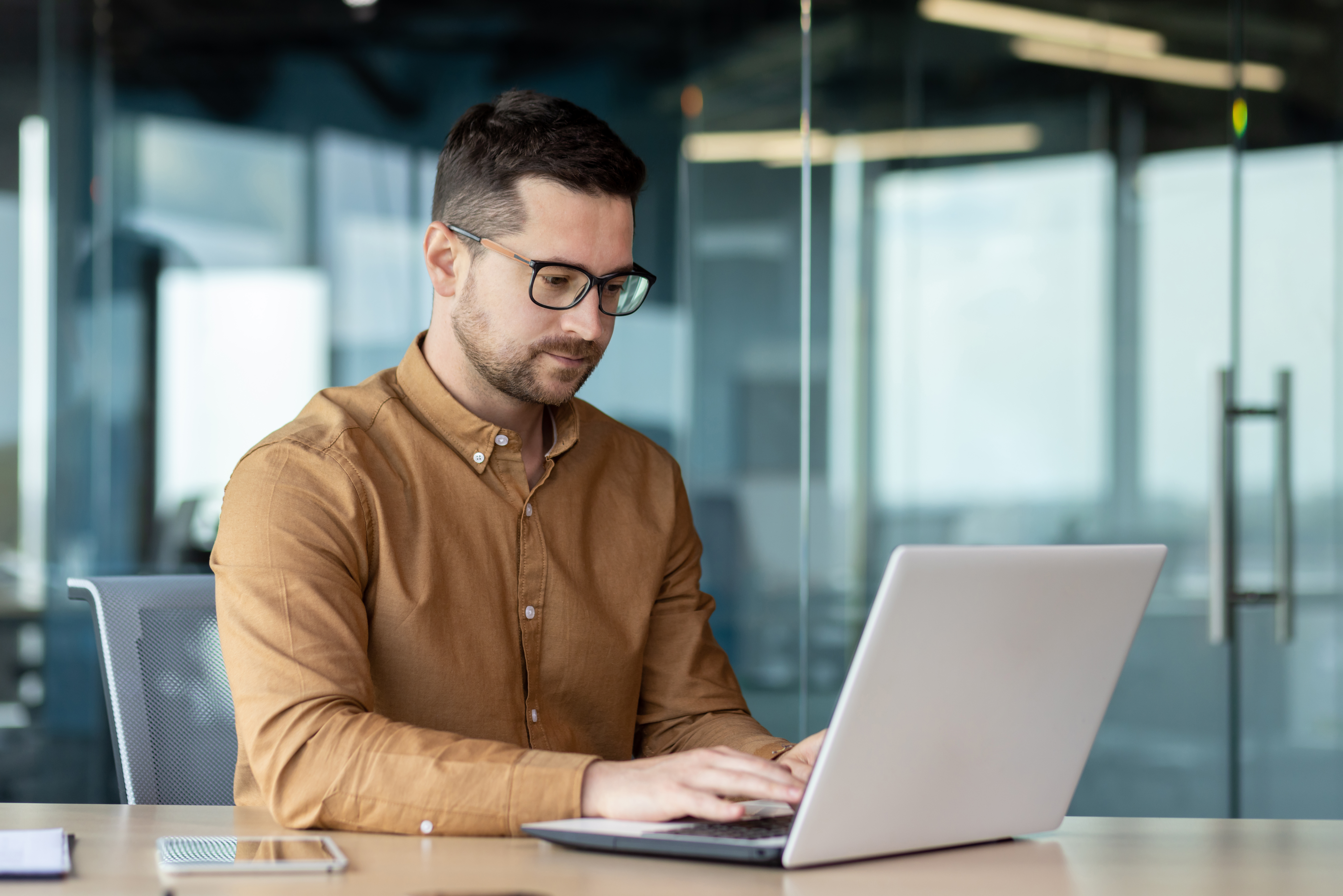Portrait of a young male programmer, in a brown shirt and glasses who is concentrating on working on a laptop, while sitting at a desk in the office.