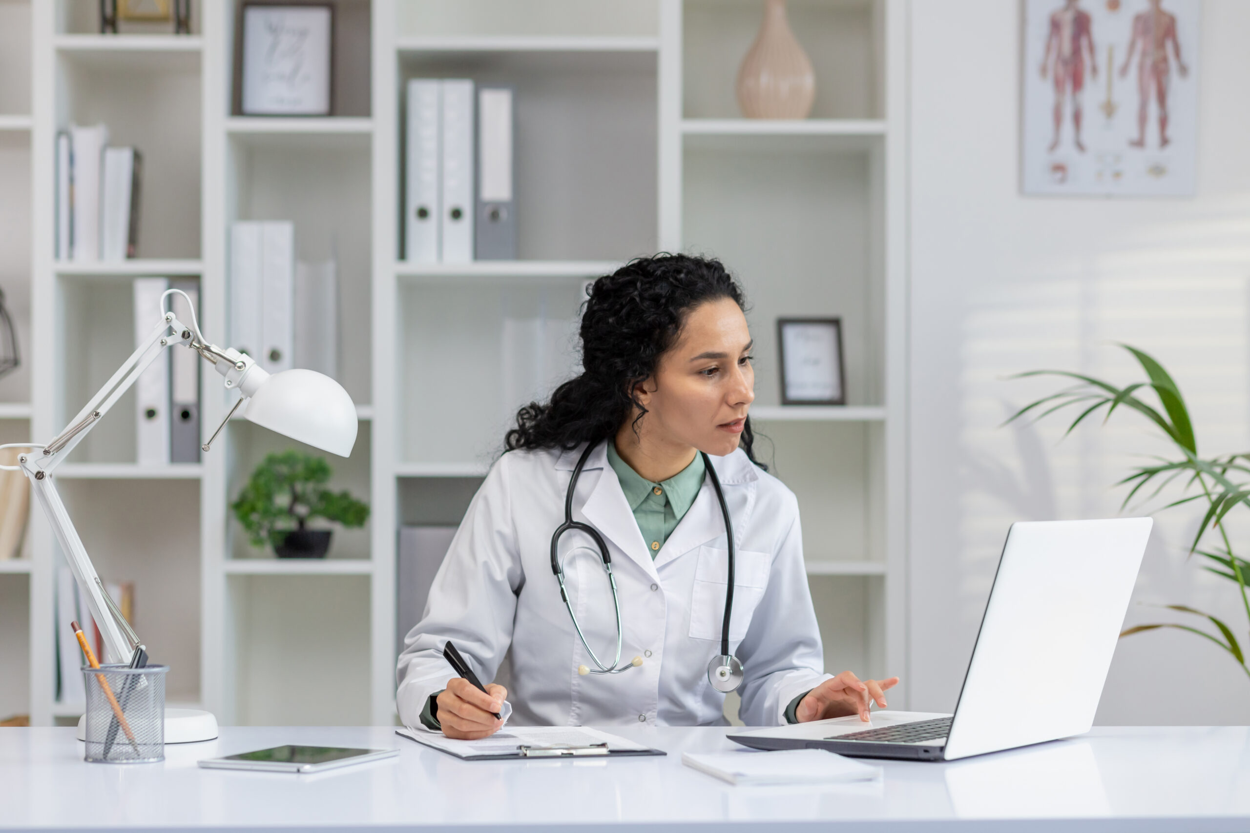 Professional hispanic female doctor in a white coat writing while working on a laptop in her office.